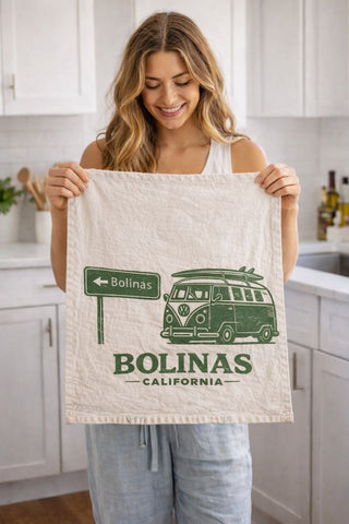 Woman holding a towel with a graphic design of a van and text that says Bolinas, California in a kitchen setting