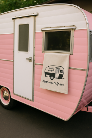 Pink vintage-style camper with a Happy Camper Petaluma California towel hanging on the door, featuring text and graphics.