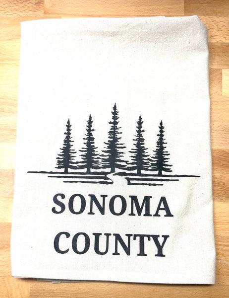 Natural color organic flour sack tea towel with black tree design and 'Sonoma County' text on a wooden surface